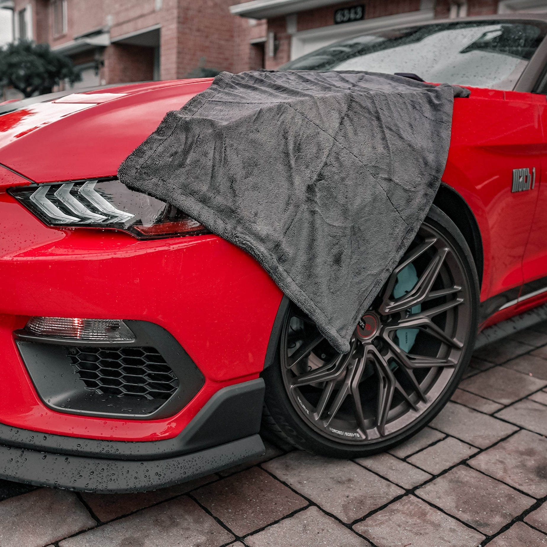 Red car with a black car drying microfiber towel draped over the hood on a brick driveway.