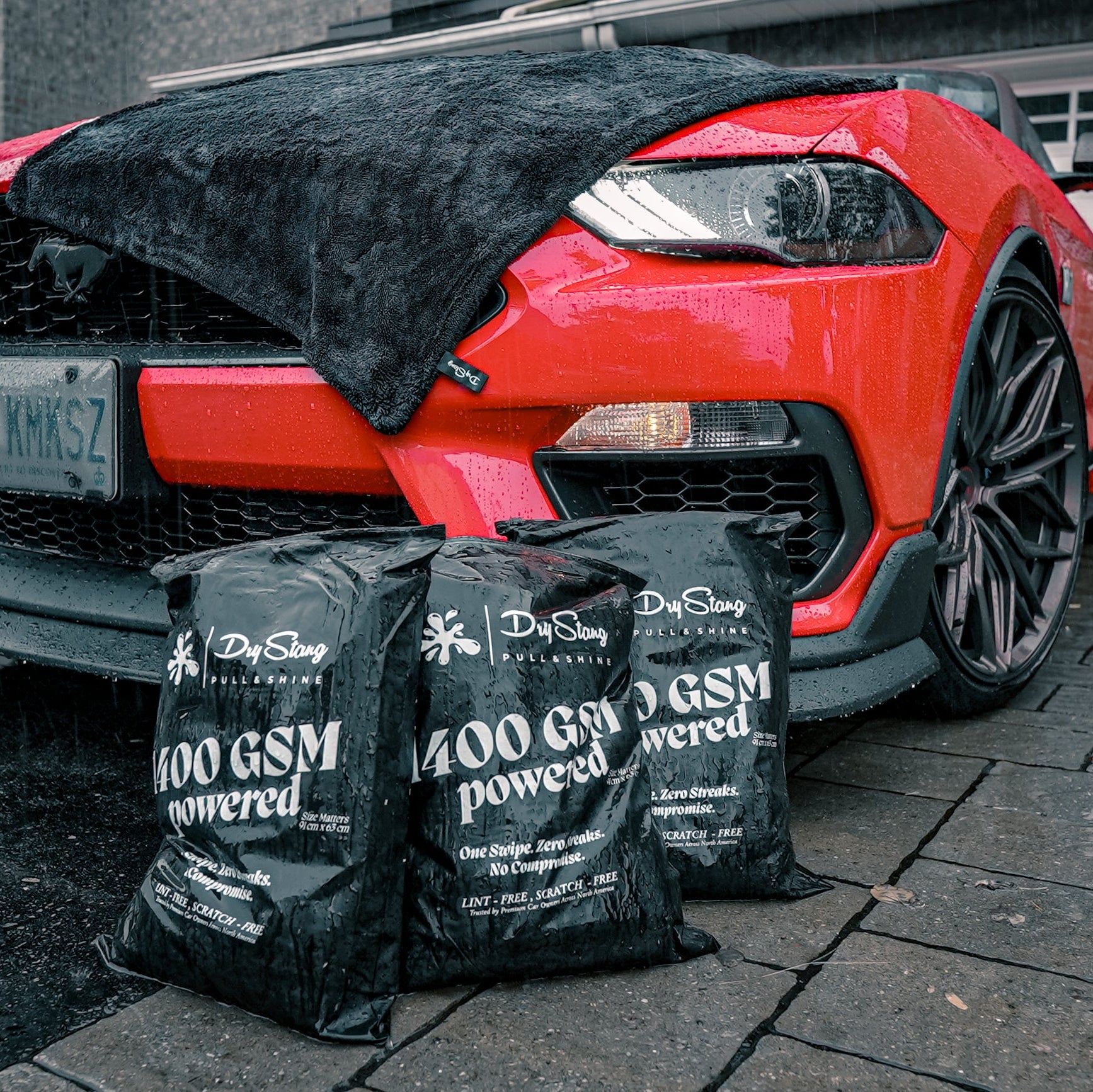 Red car with a black towel draped over it, surrounded by '1400 GSM' bags on a wet driveway.